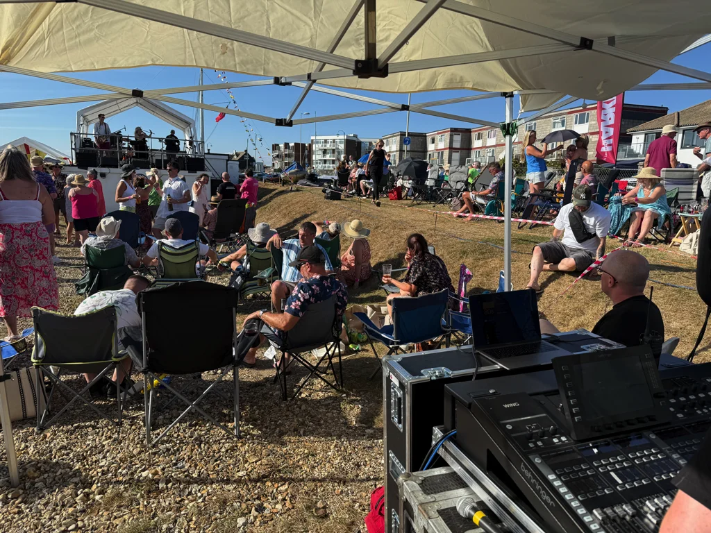 Outdoor live music event on a sunny day with people sitting on camping chairs under gazebos, while a band performs on a stage in the background and sound equipment is operated from a covered control area