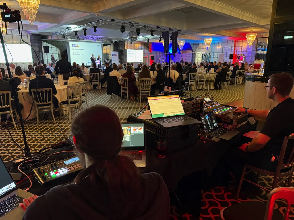 AV control team operating video, audio and lighting equipment at the back of a large banquet‑style event, with delegates seated at round tables watching presentations on stage under colourful venue lighting