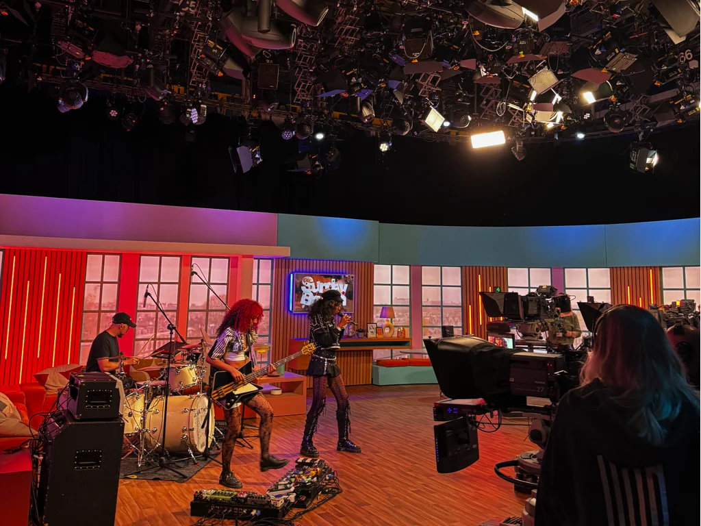 Live band performing on a brightly lit television studio set with drums, guitars, and multiple cameras filming under an extensive overhead lighting rig.